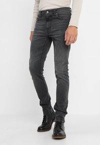 Šedé džíny z denimu se štíhlým střihem, pěticípým stylem a kontrastním šitím. Nosené s černými šněrovacími botami. Hladká textura.