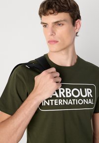 Olivově zelené bavlněné tričko s bílým textem "HARBOUR INTERNATIONAL". Model drží černý popruh, čímž předvádí jednoduchý, ležérní design.