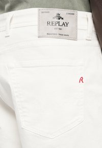 Fehér farmernadrág, amelyen a "REPLAY" márkacímke található, a hátsó zsebében egy varrt piros "R" díszítéssel, sima textúrával.