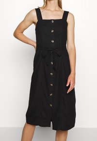 Svart ärmlös klänning med fyrkantig halsringning, knappar framtill, ett knytband i midjan och en midilängd. Tillverkad av textil med struktur.