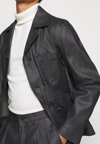 Mies, der einen dunkelgrauen Denim-Sakko und eine passende Hose mit einem weißen Rollkragenpullover trägt, mit der Hand in der Tasche, die leicht nach rechts schaut.
