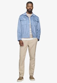 Mężczyzna stojący z jedną ręką w kieszeni, ubrany w jasnoniebieską kurtkę, beżowy sweter w paski, spodnie khaki i brązowe buty wsuwane.