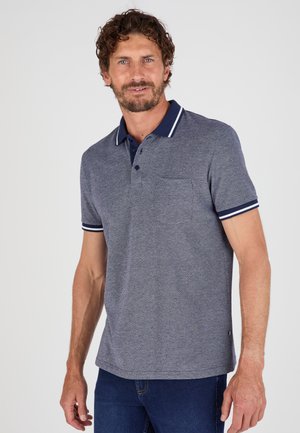 Uomo con capelli ricci che indossa una polo grigia a maniche corte con colletto blu navy e polsini delle maniche a righe, abbinata a jeans blu scuro.