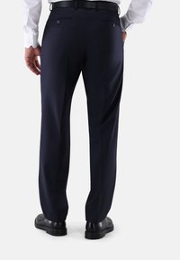 Mann, der dunkelblaue Anzughose, einen schwarzen Gürtel, schwarze Schuhe und ein weißes Hemd trägt, von hinten vor einem weißen Hintergrund zu sehen.