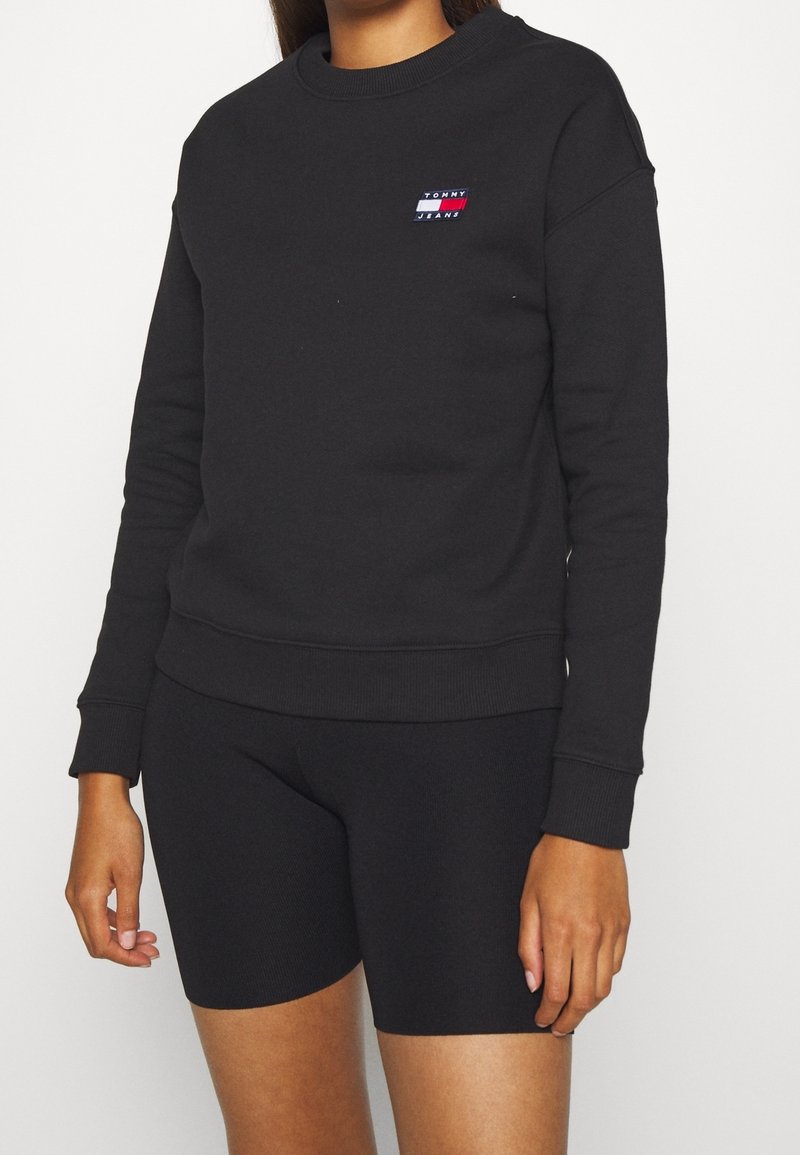 Femme portant un sweat-shirt noir à manches longues avec un petit logo Tommy Jeans et un short noir ajusté sur un fond uni.