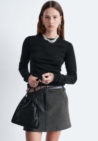 Schwarzes Langarm-Top mit gerippten Bündchen, grauer Tweedrock mit Taschen und kleine schwarze Beuteltasche mit Kordelzug. Silberne Kettenkette und Ringschmuck.