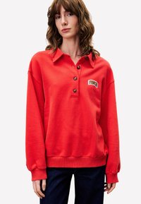 Pull rouge avec col, trois boutons et logo brodé. Équipé de poignets et d'ourlet côtelés, fabriqué en tissu doux.