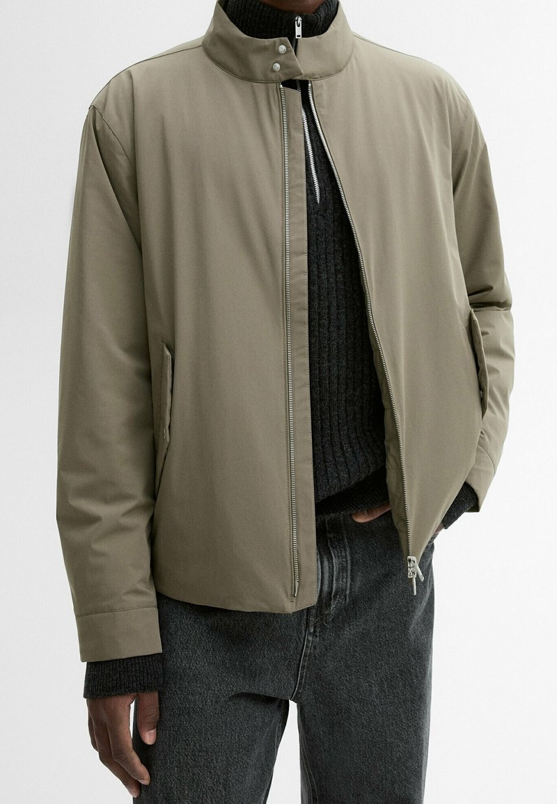 Homme portant une veste marron clair zippée par-dessus un pull noir côtelé et un jean gris foncé, une main dans la poche.