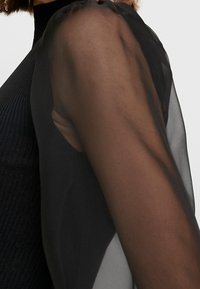 Top nero con corpo a costine e maniche trasparenti, leggere e drappeggiate con un lieve gonfiore alle spalle, creando un effetto stratificato.