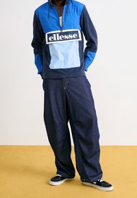Persona che indossa una felpa ellesse blu e navy con tasca frontale, jeans scuri larghi e scarpe nere e bianche su un pavimento giallo.