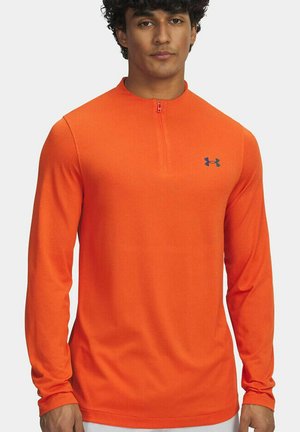 Mannelijk model draagt een feloranje sportshirt met lange mouwen, een korte ritssluiting aan de voorkant en een klein Under Armour-logo op de borst.