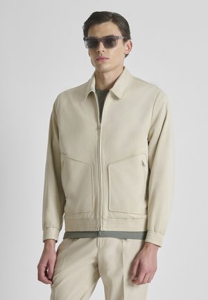 Man draagt beige jas met rits, bijpassende broek, donkergroen overhemd daaronder en donkere rechthoekige zonnebril tegen een effen achtergrond.