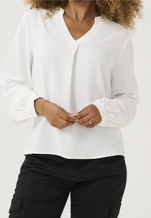 Vrouw die een witte blouse met lange mouwen met knoopmanchetten draagt, zwarte broek, gouden kettingkettingen en een ring aan haar linkerhand.