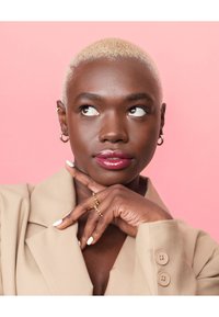 Beige blazer with large lapels, short blonde hair, dark skin, glossy pink lips, ring on finger, white manicure, and gold earrings. Pink background.