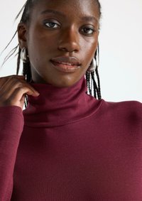 Maglione a collo alto ribbed color bordeaux con maniche lunghe, design aderente, texture morbida e alto colletto, accompagnato da una finitura liscia sulla pelle.