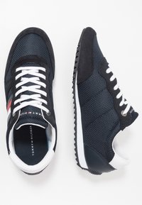 Sneakers na sznurówki w granatowym, siateczkowym materiale z czarnymi zamszowymi akcentami, biała gumowa podeszwa oraz czerwono-białe paski po boku.