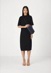 Kobieta w czarnej sukience midi trzymająca granatową i czarną torebkę na ramię, w beżowych sandałkach na obcasie, stojąca na białym tle.