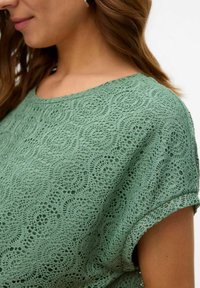 Top de encaje verde con escote redondo, mangas cortas y un patrón texturizado que presenta remolinos y motivos florales. Acabado de tela suave.