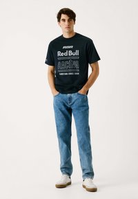 Camiseta de algodón negra con el logo de "Red Bull Racing" y texto blanco, combinada con jeans de mezclilla azul claro y zapatillas blancas con suelas marrones.
