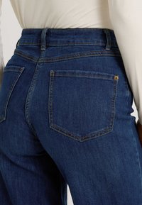 Jeans de mezclilla azul de cintura alta vistos desde la parte trasera, con dos bolsillos traseros y costuras en contraste a lo largo de las costuras.