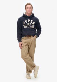 Sudadera azul oscura con el texto "SUPER" en crema claro. Pantalones caqui y zapatillas ligeras. Corte casual con capucha ajustable y bolsillo frontal.