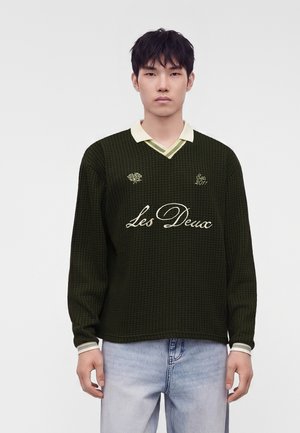 Ung mand med kort sort hår iført en mørkegrøn sweater med teksten "Les Deux" og lyseblå jeans, stående foran en ensfarvet hvid baggrund.