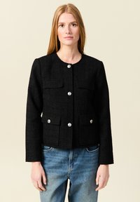 Schwarze Tweedjacke mit rundem Halsausschnitt, vorderer Knopfverschluss und zwei Brusttaschen. Mit silberfarbenen Knöpfen und langen Ärmeln.