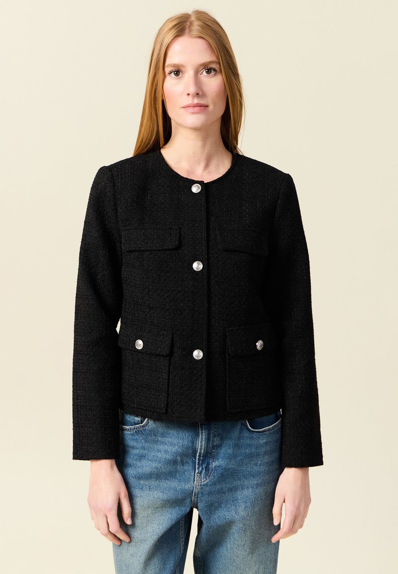 Schwarze Tweedjacke mit rundem Halsausschnitt, vorderer Knopfverschluss und zwei Brusttaschen. Mit silberfarbenen Knöpfen und langen Ärmeln.