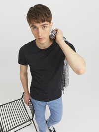 Schwarzes Baumwoll-T-Shirt, kurzärmelig, Rundhalsausschnitt. Helle Jeans aus Denim, schwarze Sneakers, ein kariertes Hemd tragend. Minimalistische Umgebung.