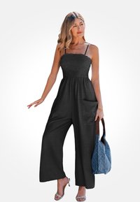 Zwarte, mouwloze jumpsuit met een geschulpt lijfje en een ontwerp met wijde pijpen; inclusief zijzakken. Gecombineerd met een gestructureerde blauwe tas.