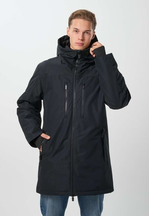 Jeune homme portant un manteau d'hiver noir à capuche avec des poches zippées, debout, une main dans la poche et l'autre ajustant la capuche.