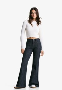 Pulover alb cu nervuri, scurt, și blugi evazați din denim închis. Blugii au talie înaltă și două buzunare pe spate. Adidașii albi completează ținuta.