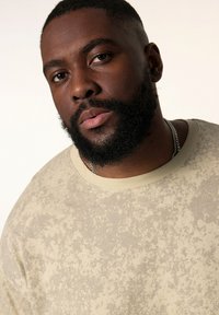 Beige sweatshirt med struktureret mønster, rund halsudskæring og ribbet kant. En sølvfarvet halskæde er synlig på emnet.