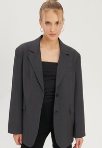 Frau mit blonden Haaren zu einem Dutt gebunden, trägt Creolen, ein graues, übergroßes Blazer, ein schwarzes Trägertop und schwarze Hose, steht vor einem schlichten Hintergrund.