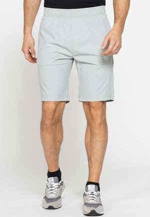 Uomo che indossa pantaloncini grigio chiaro fino al ginocchio, calzini neri alla caviglia e scarpe da corsa grigie, in piedi su uno sfondo uniforme.
