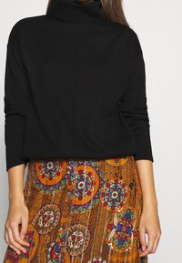 Mujer vestida con un suéter negro de cuello alto y una falda de patrón multicolor con diseños circulares y florales.