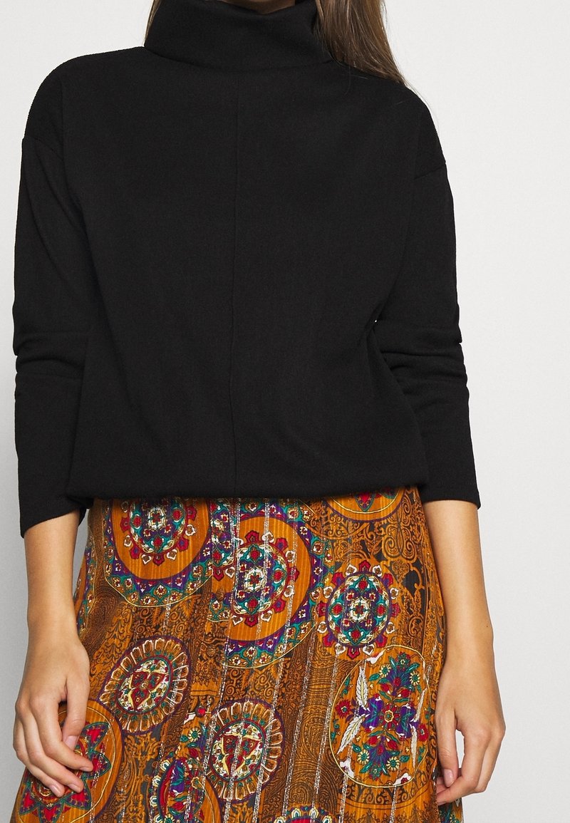Mujer vestida con un suéter negro de cuello alto y una falda de patrón multicolor con diseños circulares y florales.