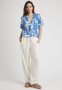 Chemise à manches courtes bleu et blanche à motifs, associée à un pantalon large crème et des espadrilles compensées beige. Fond blanc.
