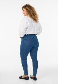 Jeans de mezclilla azul de cintura alta con un diseño ajustado y bolsillos traseros, combinados con una blusa blanca holgada y bailarinas negras.