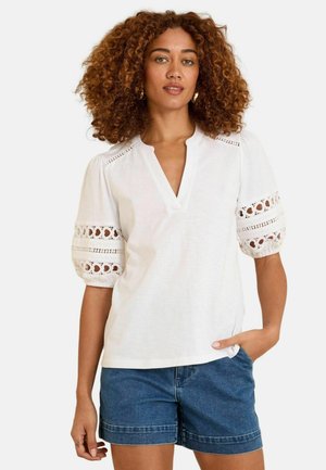 Frau mit lockigem Haar, die eine weiße Bluse mit Spitzen-Puffärmeln und blaue Jeansshorts trägt und mit einer Hand in der Tasche steht.