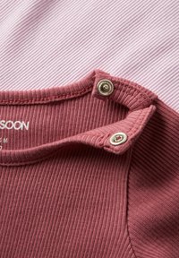 Primo piano di una scollatura in tessuto rosa a coste con due bottoni a pressione argentati su un indumento per bambini, su uno sfondo a righe rosa e bianco.