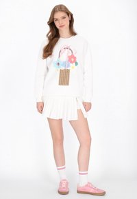 Fehér pulóver színes virágkosár hímzéssel, fehér pliszírozott szoknyával párosítva. A modell rózsaszín sportcipőt és csíkos fehér zoknit visel.
