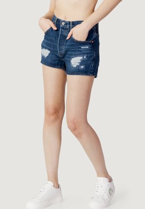Pantaloni corti in denim blu con un design strappato, orlo sfrangiato e tasche frontali, abbinati a sneaker bianche, che mostrano un look casual.