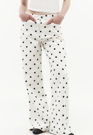 Personne portant un pantalon blanc taille haute à pois noirs, les mains dans les poches avant, associé à des chaussures noires et un top court blanc.