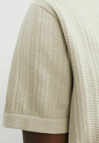 Ljust beige stickad kortärmad skjorta med ett strukturerat mönster, ribbade muddar och en mjuk yta. Visar tygets detaljer och sömnad.