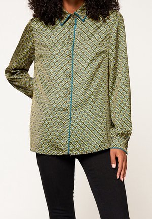 Vrouw die een groen overhemd met blauw ruitpatroon en zwarte broek draagt, staand met één hand op de heup tegen een witte achtergrond.