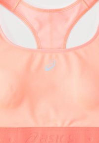 Ροδακινί αθλητικό σουτιέν με λείο ύφασμα, λεπτομέρειες από δίχτυ και φαρδιά ταινία. Διαθέτει αντανακλαστικό λογότυπο και σχέδιο racerback για υποστήριξη και διαπνοή.