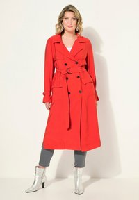 Rode trenchcoat met een dubbelzijdige sluiting, lange mouwen en een riem in de taille. Voorzien van twee voorkruiszakken en zijsplitten. Draagt over grijze broek.