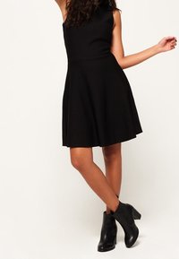 Robe noire sans manches, ajustée à la taille et évasée, en tissu texturé, avec une taille cintrée et une jupe évasée. Associée à des bottes noires à chevilles.