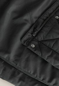 Giacca nera trapuntata con finitura lucida, chiusura con zip e dettagli con bottoni a pressione in un design elegante. Tessuto morbido e leggero.